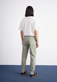 Cămașă scurtă gri deschis, combinată cu pantaloni verzi măslinii. Modelul stă de profil pentru a arăta cum se potrivește. Încălțăminte texturată închisă la culoare cu talpă din cauciuc.