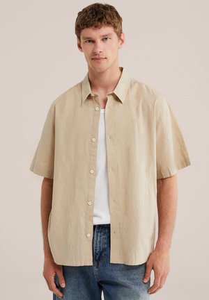 Jeune homme aux cheveux bruns courts, portant une chemise beige à manches courtes boutonnée sur un t-shirt blanc et un jean bleu, debout devant un fond uni.