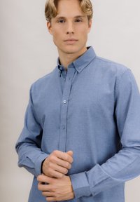 Lyseblå skjorte med knapper, laget av mykt stoff, med krage, lange ermer og knappedetaljer foran og på mansjettene.