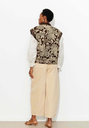 Femme aux cheveux courts portant un gilet marron à motif floral, un chemisier blanc à manches longues en broderie anglaise, un pantalon large beige et des sandales marron à talons.