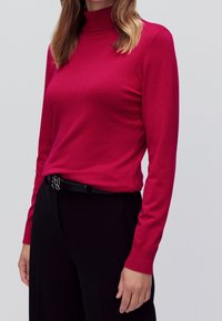 Magenta Rollkragenoberteil aus glattem Stoff, mit langen Ärmeln und enganliegendem Schnitt, kombiniert mit hochgeschnittenen schwarzen Hosen und einem Gürtel.