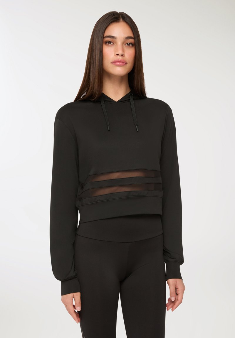 Donna con capelli lunghi e lisci che indossa un hoodie nero a maniche corte con pannelli orizzontali trasparenti e leggings neri, in piedi davanti a uno sfondo semplice.