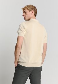 Crème korte mouwen polo shirt, geribde manchetten en onderkant, zachte gebreide stof, casual pasvorm, gecombineerd met donkere broek, gezien van achteren.