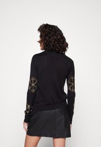 Schwarzer Pullover mit langen Ärmeln, der goldene Stickereien im Schneeflockenmuster auf den Ärmeln aufweist, aus einem weichen, strukturierten Material gefertigt.