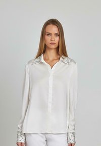 Blusa bianca in satin con collare e polsini decorati da applicazioni brillanti. Maniche lunghe con chiusura frontale a bottoni. Dettaglio di texture del tessuto liscio.