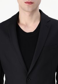 Zwarte blazer met notch-revers en een enkele knoopsluiting, gedragen over een gestructureerde zwarte trui. Gladde stof met een getailleerde pasvorm en zakdetails.