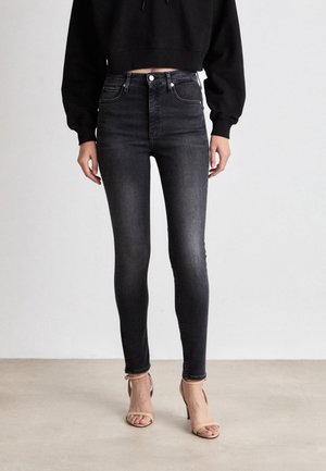 Femme portant un jean skinny noir délavé taille haute, un sweat-shirt court noir et des sandales beiges à talons hauts, debout sur un sol uni.