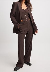 Brown op maat gemaakt pak met een blazer met gelapte revers, een kort knoopshirt met zilveren knopen en rechte broekspijpen. Gladde stofstructuur.