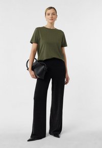 Kobieta stoi w oliwkowej, krótkofalowej bluzce, czarnych, szerokich spodniach i czarnych, szpiczastych butach, trzymając czarną torebkę przy boku.