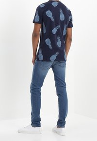 Camisa azul marino de manga corta con estampado de piñas en azul claro, combinada con vaqueros azul claro y zapatillas blancas. Diseño simple y casual.