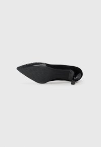 Black high-heeled shoe made of suede, with a pointed toe, textured sole, and a decorative strap at the back. Detailed with small accents.