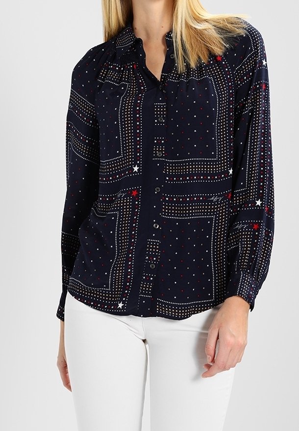 Femme portant un chemisier bleu marine à boutons avec des motifs géométriques et d'étoiles, associée à un pantalon blanc, debout devant un fond uni.