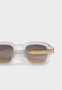 Διαφανή γκρι γυαλιά ηλίου με ορθογώνιους φακούς, καφέ gradient σκίαση και χρυσές λεπτομέρειες στα γέφυρα που περιλαμβάνουν ένα λογότυπο.