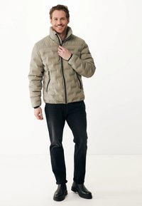 Giacca imbottita di colore verde oliva con colletto alto e chiusura con zip, dotata di cerniere nere a contrasto. Indossata con jeans scuri e stivali neri.