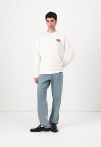 Kremgenser med rød logo-patch, sammen med lyseblå løstsittende denimjeans og svarte lærsko, som står mot en hvit bakgrunn.