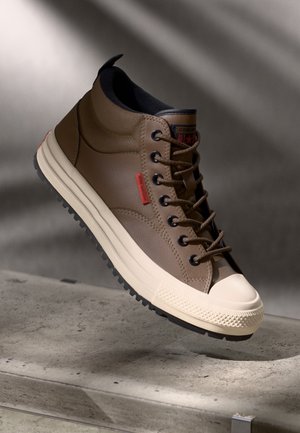 Brown high-top sneaker with cream rubber toe cap and sole, black laces, and red tag detail, positioned above a concrete surface.