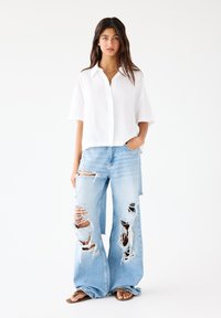 White short-sleeve button-up shirt paired with light blue distressed wide-leg jeans featuring large rips and frayed edges, worn with brown sandals.