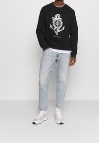 Sweat-shirt noir avec graphisme d'ancre nautique, jean bleu clair et baskets grises. Le sweat-shirt présente des poignets et un ourlet côtelés.