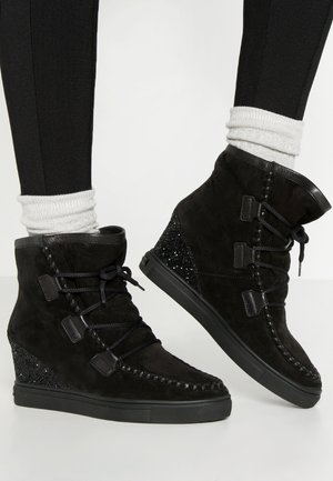 Bottines noires à semelle compensée avec des accents scintillants au talon, lacées à l'avant, portées avec des chaussettes blanches et des leggings noirs.