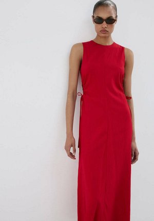 Femme portant une robe rouge sans manches avec des liens sur les côtés et des lunettes de soleil noires, debout devant un fond blanc uni.