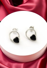 Silverfärgade hoopörhängen med svarta hjärtformade stenar. Slät yta, minimalistisk design och säker låsning. Visas på en vit ställ.