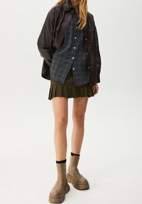 Mörkbrun läderjacka, blå och grön rutmönstrad skjorta med knappar, olivgrön plisserad minikjol och beige plattformskängor med svarta detaljer.