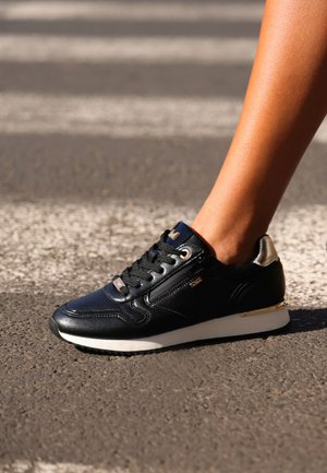Zwarte leren sneaker met gouden accenten, ronde neus, vetersluiting, gestructureerd oppervlak en een witte zool, geplaatst op een zebrapad.