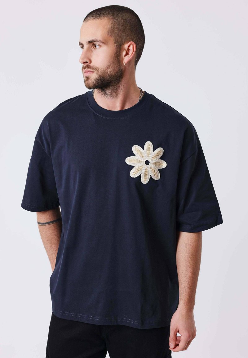 Homme aux cheveux courts et barbe, portant un t-shirt bleu marine avec un grand motif de fleur crème sur la poitrine, regardant vers la gauche.