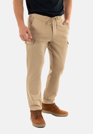 Pantalons cargo beiges avec une taille à cordon, des poches latérales et un tissu texturé. Associés à des chaussures brunes à lacets pour un look décontracté.