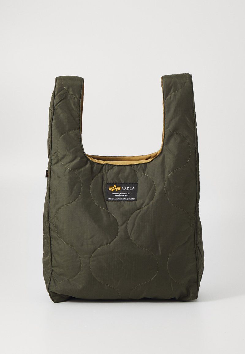 Sac fourre-tout vert matelassé de forme arrondie, avec un intérieur beige et un patch logo à l'avant. La texture est lisse et moelleuse.