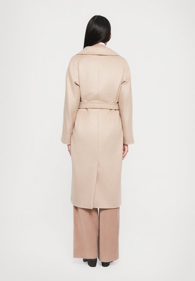 Long, belted coat in light beige, made of textured fabric. Features a notched collar and a vent at the back. Worn with pleated pants.