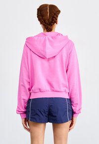 Cropped hoodie rosa brillante con cappuccio regolabile, tessuto morbido e polsini a costine, abbinato a pantaloni corti blu scuro con una striscia laterale bianca.