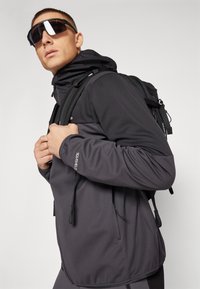 Jachetă softshell gri închis cu glugă, dotată cu șnururi ajustabile și un logo. Purtați cu ochelari de soare negri și un rucsac.