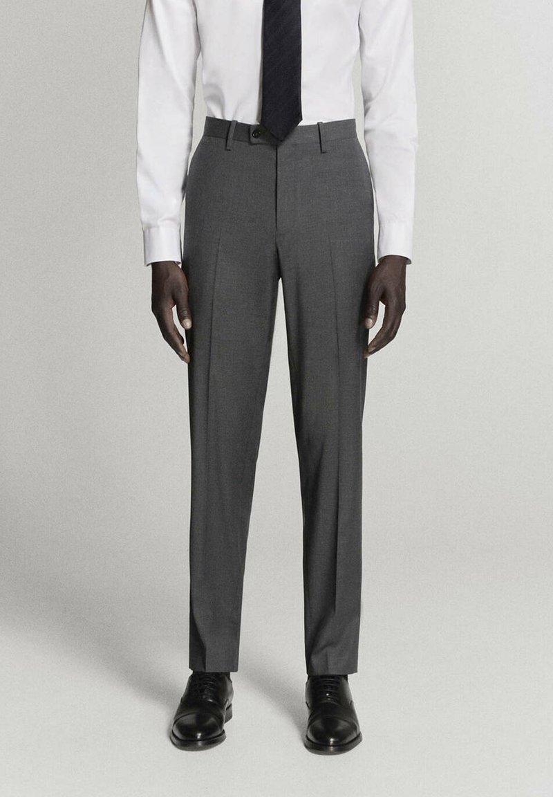 Homme portant un pantalon de costume gris avec une chemise blanche, une cravate noire et des chaussures de ville noires, debout devant un fond uni.