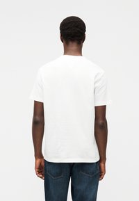 Λευκό βαμβακερό t-shirt με στρογγυλή λαιμόκοψη και κοντά μανίκια, παρουσιάζεται από πίσω. Ζευγάρι σκούρα μπλε τζιν παντελόνια με ορατές λεπτομέρειες ραφών.