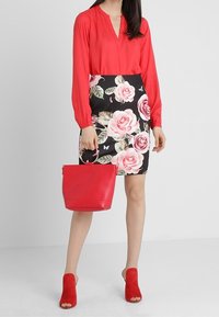 Donna che indossa una blusa rossa, una gonna nera a fiori con rose rosa, scarpe con tacco aperte rosse e tiene in mano una borsa rossa su uno sfondo semplice.