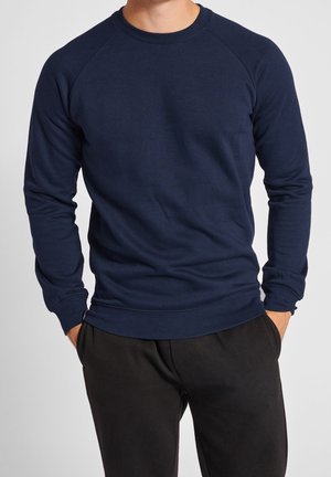 Mand iført en tætsiddende marineblå langærmet sweatshirt med raglanærmer og sorte bukser, hænderne i lommerne, mod en ensfarvet baggrund.