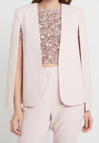 Vaaleanpunainen bleiseri avoimella edustalla, jossa on viitta-tyyliset hihat. Alla samansävyinen, paljettein koristeltu crop top. Pehmeä, sileä pinta.