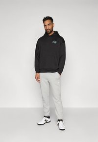 Fekete kapucnis pulóver kék "City" hímzéssel, szürke melegítőnadrággal és fehér sportcipővel párosítva. Simább anyag, laza szabás.