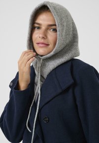 Mujer vistiendo un abrigo azul marino y una capucha de punto gris con cordones, tocando la capucha cerca de su rostro, frente a un fondo liso.