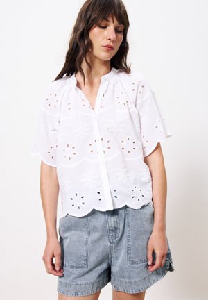 Blouse en coton blanc avec des motifs floraux brodés, ourlet festonné, manches courtes et devant boutonné, associée à un short en denim clair.