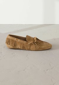 Scarpa mocassino in suede marrone con dettagli in frange, cuciture decorative e grommet in metallo. Suola piatta con design della punta arrotondata.