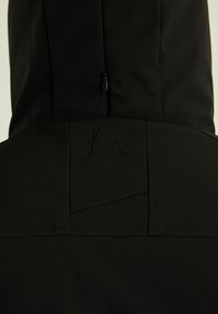 Vue arrière d'une veste noire avec un logo cousu, des détails de couture, et une courte fermeture éclair sur la capuche.