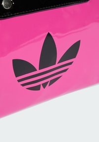 Rózsaszín fényes táska fekete Adidas logóval, amely három levelet és három vízszintes csíkot ábrázol. Fekete díszítés és patentos zárás.