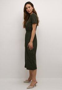 Olivgrünes, knielanges Kleid mit kurzen Ärmeln, elastischem Bund und glatter Textur. Ein Paar beigefarbene, mit Absätzen versehene Mules rundet das Outfit ab.