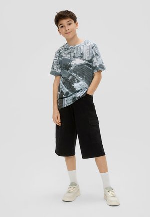 Grijze en zwarte graphic t-shirt met een stadssilhouet ontwerp, gecombineerd met losse, zwarte cargo shorts. Schoeisel bestaat uit crèmekleurige sneakers en witte sokken.