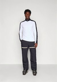 Vyras stovi vilkėdamas baltą ir juodą ilgomis rankovėmis užtraukiamą viršutinią dalį, juodas kelnes su smėlio spalvos užtrauktukais ir juodus slidinėjimo batus prieš paprastą foną.