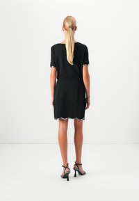 Femme avec une longue queue de cheval blonde, tournée le dos, portant une robe noire avec un ourlet en dentelle et des escarpins noirs à talons hauts.