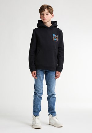 Hoodie noir avec un design graphique featuring des formes bleues, brunes et beiges. Associé à un jean en denim bleu clair et à des baskets blanches.