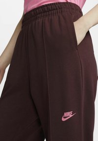 Burgundy sweatpants with elastic waistband, side pockets, and pink Nike logo on the left thigh. Soft, smooth texture.